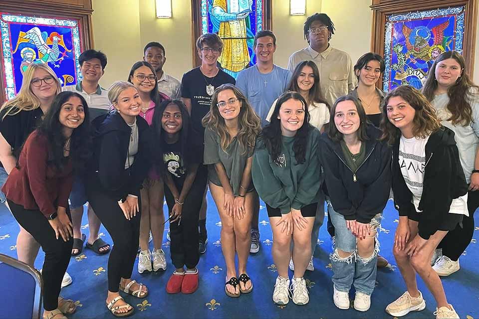 A group of 17 students pose for a photo together in a room with stained glass windows behind them.